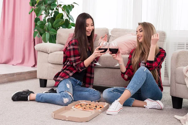 Il est temps de se détendre ! Deux filles élégantes et élégantes habillées de jeans bleus avec chemises à carreaux rouges cliquetis verres avec du vin pour un toast tout en étant assis sur le tapis à la maison . — Photo