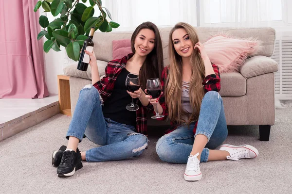 Bucură-te de viaţa ta! Două tinere zâmbind doamne prețioase așezate pe podea, bând vin roșu și pozând la camera foto într-o atmosferă confortabilă . — Fotografie, imagine de stoc
