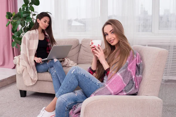 Il est temps de se détendre ! Deux belles filles rêveuses assises sur le canapé. L'un d'eux surfe sur Internet et un autre boit une tasse de café riche en s'amusant et en souriant à la caméra . — Photo