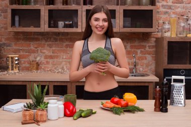 Sağlıklı bir kalp için akıllı yemek! Onun önünde büyük bir taze brokoli tutmak ve mutfakta fotoğraf için poz uygun olmak sunan güzel atletik gülümseyen kız.