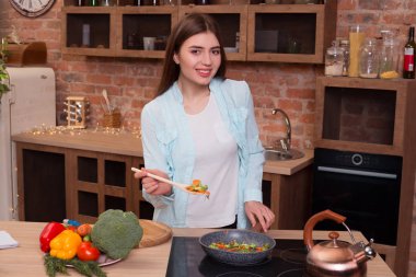 Yemek değil, parlak! Güzel sportif genç kız kızartma sebze ve kameraya gülümseyen yemek kaşığı kadar holding modern mutfak pişirme.