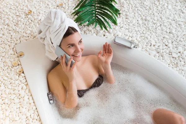 Bringing the future of healthcare! Attractive charming lady having the rest in the bath with a towel on her head and enthusiastically talking her phone.