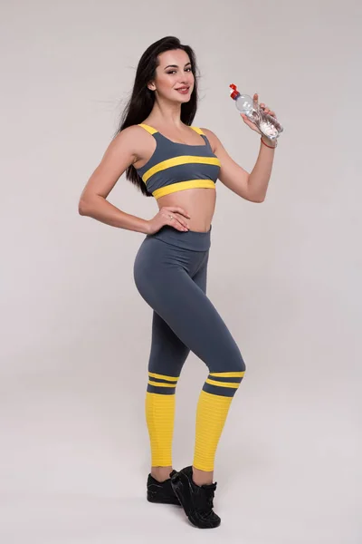 Ingenting vil fungere med mindre du gjør det! Sporty muskuløs ung kvinne som drikker vann, isolert mot grå bakgrunn og smiler på bildet . – stockfoto