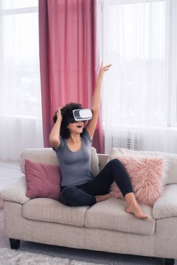 Zaman uygun şekilde pass! Muhteşem bir yakışıklı genç kadın mutlu bir şekilde gülümseyerek, konforlu gözlük sanal gerçeklik ile modern kanepede otururken.