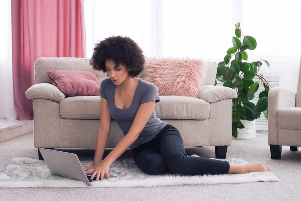 Détendez-vous le mieux possible ! Belle jolie jeune femme avec afro-coupe de cheveux assis sur le tapis mou sur le sol à la maison et tapant sur son ordinateur sans fil . — Photo