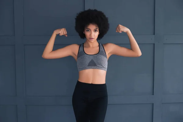 Be active, be healthy, be happy! Beautiful young strong model in good shape posing for the camera isolated on the dark grey background and demonstrating her muscles.