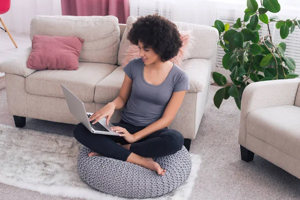 C'est le moment ! afro-américaine souriant fille joyeuse assis sur le tissu tricoté gris hassock avoir un grand temps libre avec son ordinateur portable à la maison . — Photo