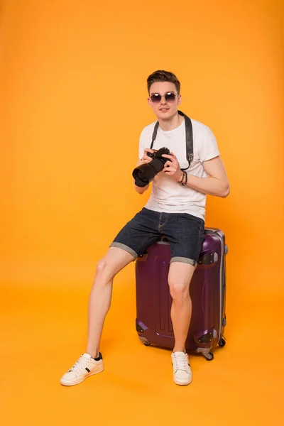 ¡Brindemos por unas vacaciones sin arrepentimientos! Retrato de cuerpo entero del inteligente hombre con gafas y cámara, sentado en la maleta . — Foto de Stock