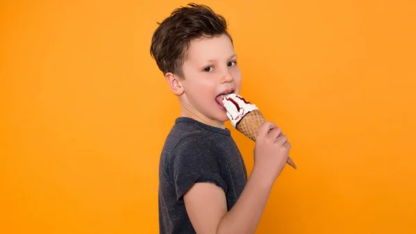 Time for yourself! Close up picture of cheerful little boy happily eating delicious ice-cream over isolated orange background.