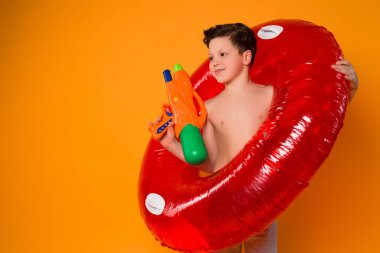 Genç iken mümkün olduğu kadar oynayın! Boya tabancası ve kauçuk halka izole turuncu arka plan üzerinde sulama ile oynayan genç çocuk gülümseyen.