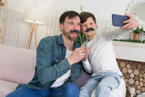 Chaque enfant grandit en pensant que son père est un héros ! Photo rapprochée du père et de son fils faisant du selfie avec une moustache, assis sur le canapé . — Photo