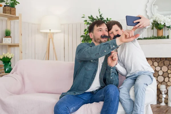 Un bon père se donne tout ce qui est bon ! Mignon fils avec son père assis sur le canapé et posant pour le selfie avec moustache . — Photo