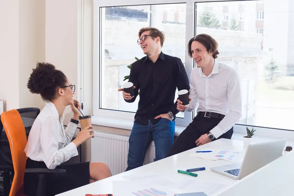 Work like you do not need the money! Smart young group of three joking and drinking coffee during the break of their work.