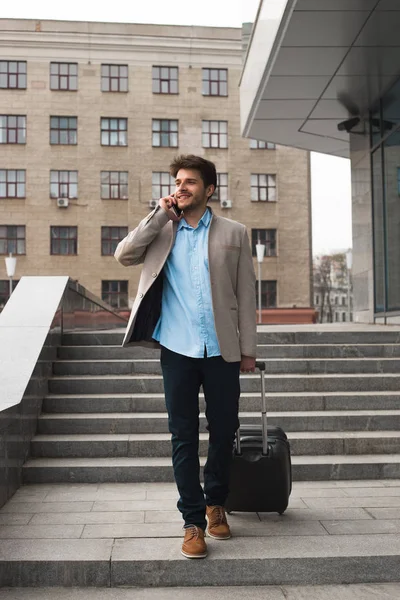 ¡Un profesional líder! Guapo joven sonriente en viaje de negocios caminando con su equipaje y hablando por teléfono celular en el aeropuerto . — Foto de Stock