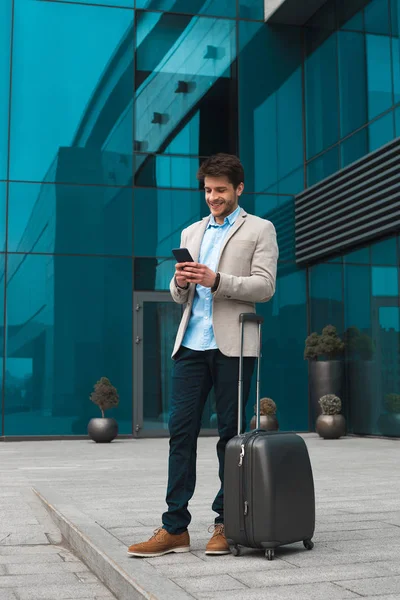 Atteindre le but ! Portrait intégral du jeune homme d'affaires élégant et intelligent qui enregistre ses e-mails sur son téléphone portable, debout près de ses bagages à l'extérieur . — Photo