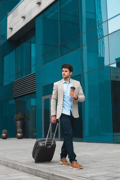 Un professionnel de premier plan ! Jeune homme d'affaires impressionnant marchant avec une valise à l'extérieur du bâtiment de l'aéroport et buvant du café à emporter . — Photo