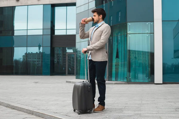 Regardez bien à ce jour ! Photo pleine longueur du bel entrepreneur délicieux buvant un délicieux café et debout avec ses bagages près de l'aéroport, en attendant le taxi . — Photo