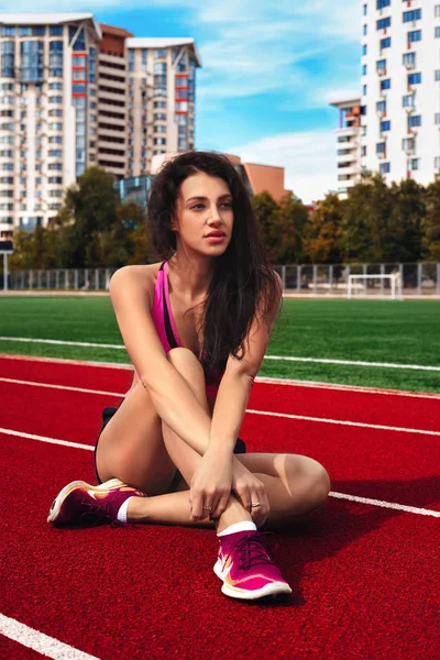 Athletic young woman sitting on a race track at a sports stadium on a bright summer day