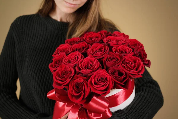 Menina de casaco preto segurando na mão rico buquê de presente de 21 rosas vermelhas. Composição de flores em um hatbox branco. Amarrado com fita vermelha larga e arco . — Fotografia de Stock