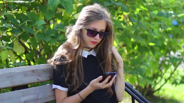 Hermosa chica en gafas de sol negras utiliza su teléfono en un banco del parque . — Vídeos de Stock