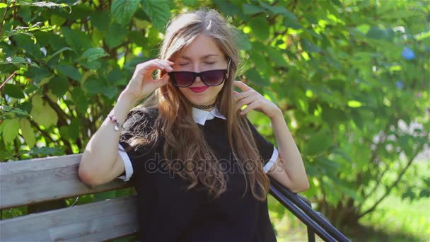 Красива дівчина в чорних сонцезахисних окулярах посміхається на лавці парку . — стокове відео