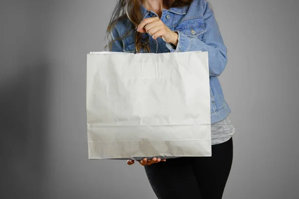 La chica en la camisa de mezclilla azul sosteniendo bolsa de papel limpia en blanco . — Foto de Stock