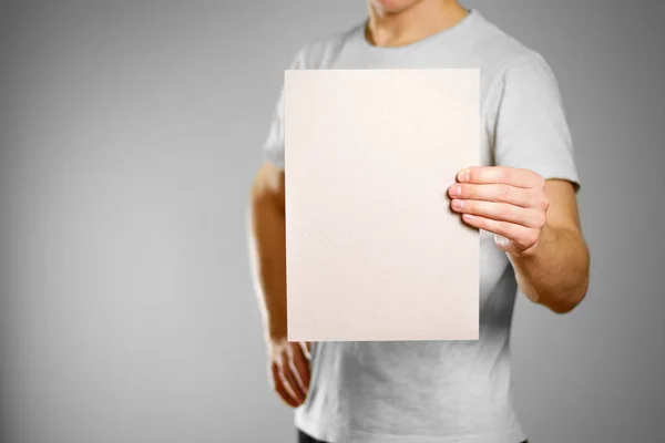 Um homem com uma t-shirt cinzenta mantém uma folha cinzenta em branco e limpa de A4. É — Fotografia de Stock