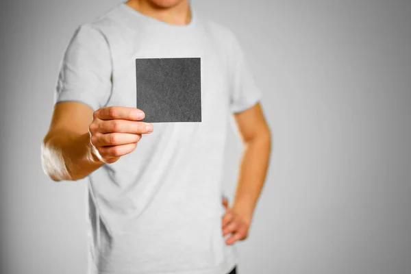 Ein Mann im grauen T-Shirt hält einen schwarzen, sauberen, leeren quadratischen Aufkleber in der Hand — Stockfoto