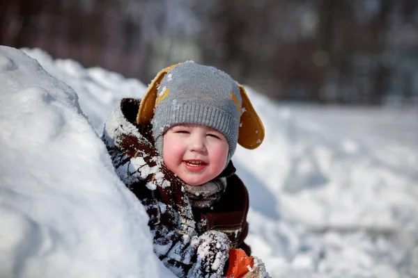 Karda güzel bir çocuğun portresi. Büyük kar yığınları var. Kulaklı gri şapka ve kahverengi kuzu derisi ceket..