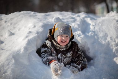 Kırmızı pantolonlu küçük yakışıklı bir çocuk ve kahverengi bir kuzu derisi ceket kar yığınları arasında yol alıyor. Çocukla oynanıyor. Arka planda kardaki ağaçlar.