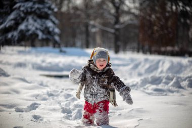 Kırmızı pantolonlu küçük yakışıklı bir çocuk ve kahverengi bir kuzu derisi ceket kar yığınları arasında yol alıyor. Çocukla oynanıyor. Arka planda kardaki ağaçlar.