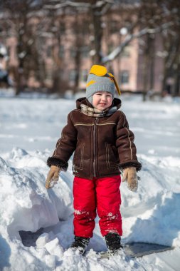 Kırmızı pantolonlu küçük yakışıklı bir çocuk ve kahverengi bir kuzu derisi ceket kar yığınları arasında yol alıyor. Çocukla oynanıyor. Arka planda kardaki ağaçlar.