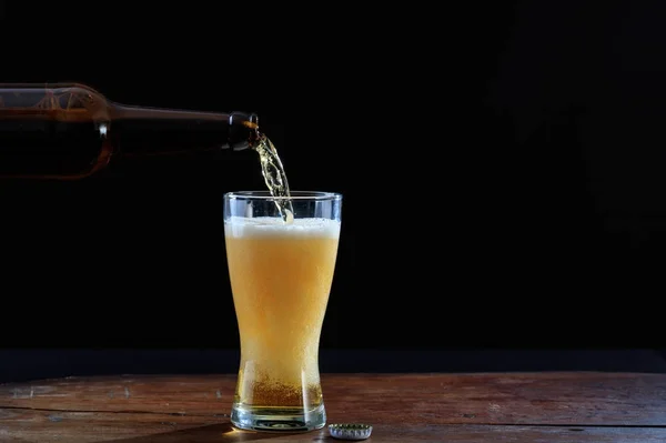 Derramando cerveja em um copo em uma mesa de madeira, fundo escuro — Fotografia de Stock