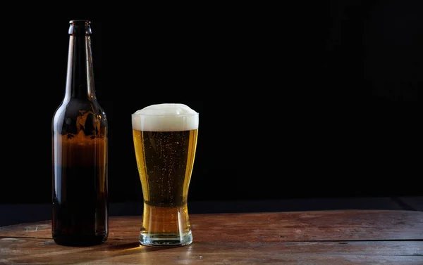 Uma garrafa e um copo de cerveja em uma mesa de madeira, fundo escuro — Fotografia de Stock