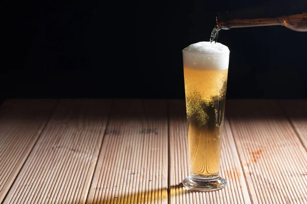 Derramando cerveja em um copo em uma mesa de madeira, fundo escuro — Fotografia de Stock