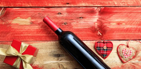 Valentines day. Red wine bottle, a gift and hearts on red wooden background — Stock Photo, Image