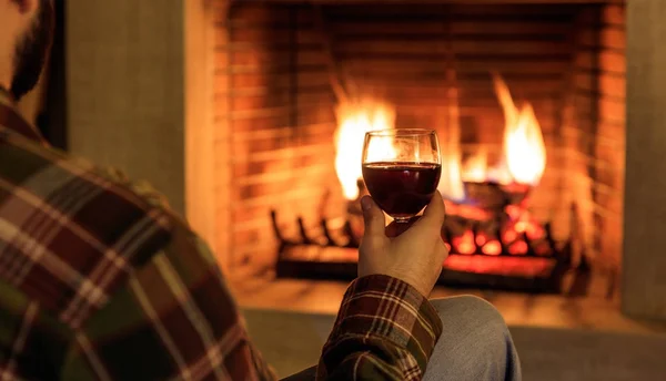 Человек держит бокал красного вина на горящем фоне камина — стоковое фото