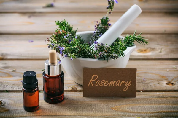 Rosemary essential oil and fresh blooming twig in a mortar, wooden table, tag with text rosemary