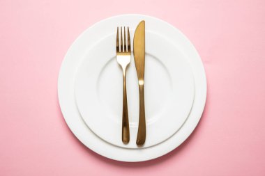 Pembe arka planda altın çatal bıçak takımı ve tabaklar.