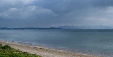 Pofuduk gri-mavi bulutlar kumlu sahilden uzakta sakin koyu mavi denizle buluşur. Dağlar arka plan, boşluk.