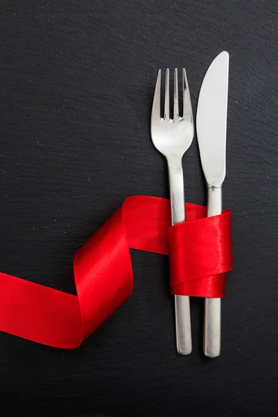 Valentýnské prostírání stolu. Vidlice a nůž příbory svázané červenou stuhou na černém pozadí, horní pohled. — Stock fotografie