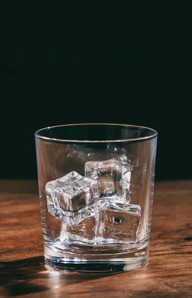 Crystal empty whiskey glass with ice cubes on wooden bar counter. Vertical portrait of a clear transparent glassware tumbler full of ice rocks on dark background.