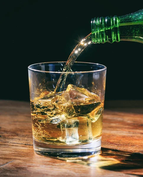 緑のボトルの岩の上に 木製のテーブルの上に結晶ガラスにウイスキーを注ぐ 暗い背景に氷のキューブとアルコール飲料の垂直肖像画 — ストック写真