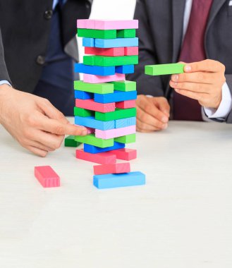 Beyaz bir ofis masasının üzerindeki renkli bloklar kulesi. Masa oyunu oynayan iki adam. İş stratejisi, risk, inşaat konsepti