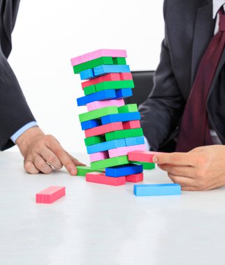 Beyaz bir ofis masasının üzerindeki renkli bloklar kulesi. Masa oyunu oynayan iki adam. İş stratejisi, risk, inşaat konsepti