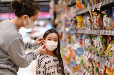 Koruyucu maske takan bir kadın süpermarketteki kızına yüz maskesi takıyor. COVID-19 salgını sırasında güvenlik. Mart 2020 'de Ramintra, Tayland' da çekilen virüs salgını..