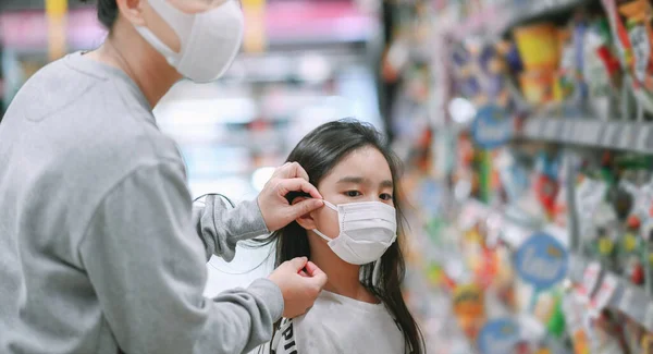 Mother wearing a protective mask puts a face mask on a daughter in  supermarket. Safety during COVID-19 outbreak. Epidemic of virus covid,