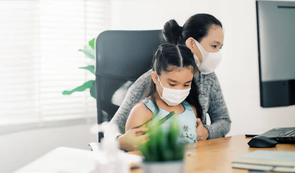 mom working at home wearing protective mask  with her child on the table while writing an report. woman working from home, while in quarantine isolation during the Covid-19 health crisis