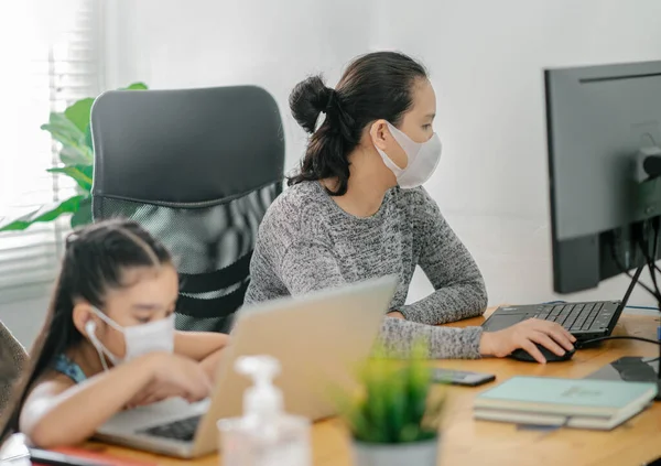 mom working at home wearing protective mask  with her child on the table while writing an report. woman working from home, while in quarantine isolation during the Covid-19 health crisis