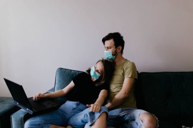 Tıbbi maskeli, birbirine aşık bir çift kanepede oturmuş dizüstü bilgisayara bakıyor. Aile Karantina Konsepti.